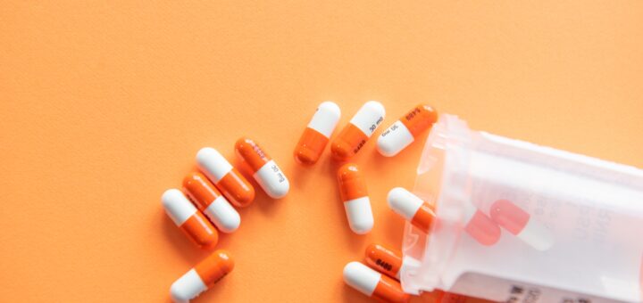 A bottle of pills is tipped sideways spilling out onto an orange background. The pills are or and white capsules.