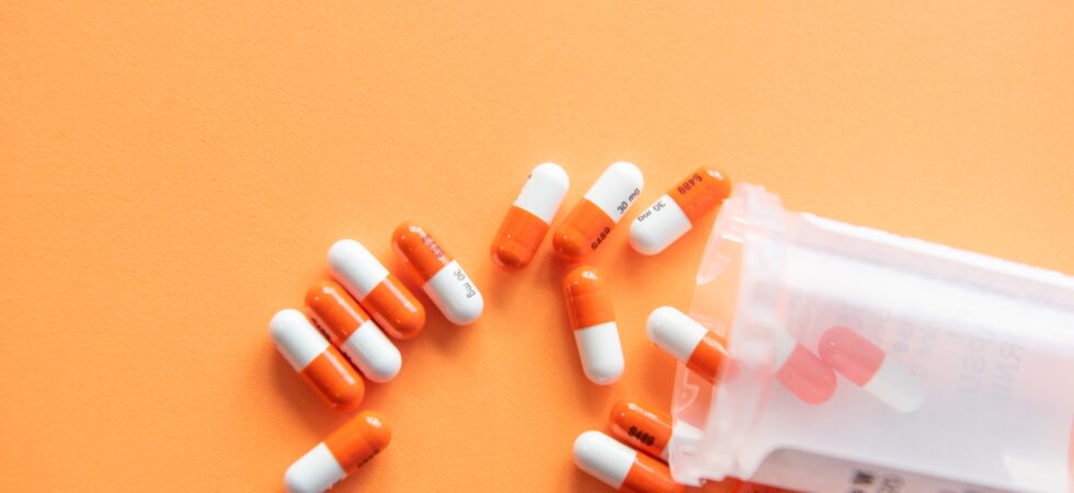 A bottle of pills is tipped sideways spilling out onto an orange background. The pills are or and white capsules.