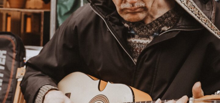 An older man in a flat pork-pie cap and brown jacket strums his in instrument in a casual setting.