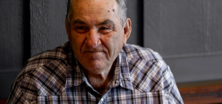 An older man in a bakc and white plaid shirt stares off to the side as is upset or angry. He wears his steel gray nd white hair combed back.
