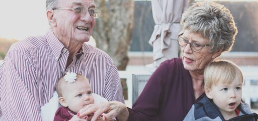 Older man and woman, likely the grandparents, holding baby and toddler on their laps. Both grandparents have gray hair. Woman is wearing a purple sweater and man is wearing a pink shirt.