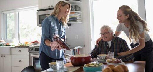 Woman in blue tee shirt standing by a table showing a book that looks like a picture album to a seated older man in a read flannel