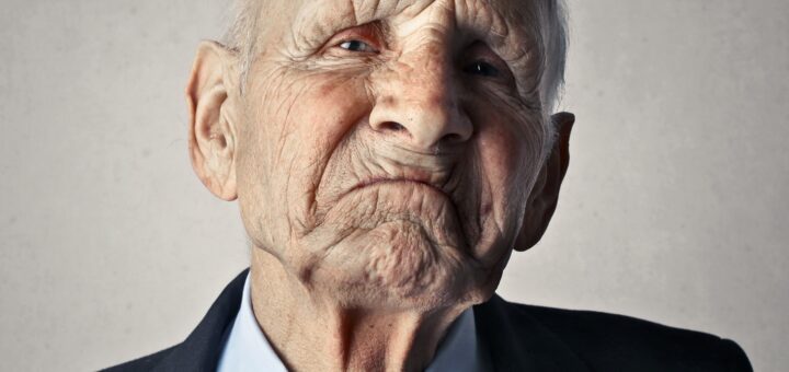 Older grandfatherly bald man in a dark suit, light blue shirt, and red tie with exagerated frown and stubborn look.