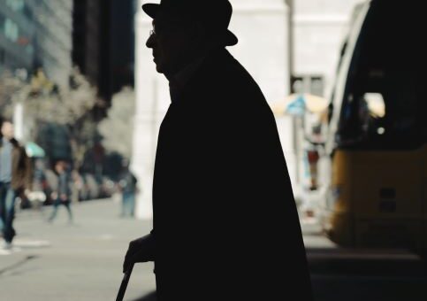 Shadow image of an older man in a bowler hat crossing a city street while using a cane