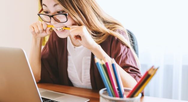 Woman with long brown hair and glasses in front of a laptop computer. Her frustration shows as she bites down on a pencil placed horizontally in her mouth.