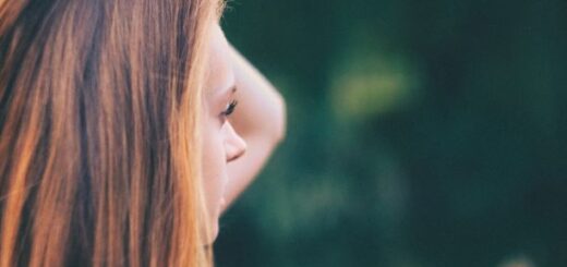 Blurred background is dark green foliage; Foreground on the lest is a woman in profile with her arm raised as if she's pushing back long brown hair. Overall image is one of emotion.