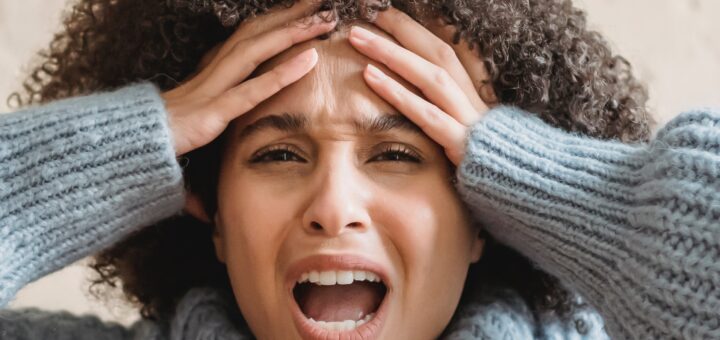 Close up of an angry woman with dark brown curly hair, wearing a chunky blue sweater. Shes got both hands on her forehead with her mouth wide open in frustration as if she's been crying
