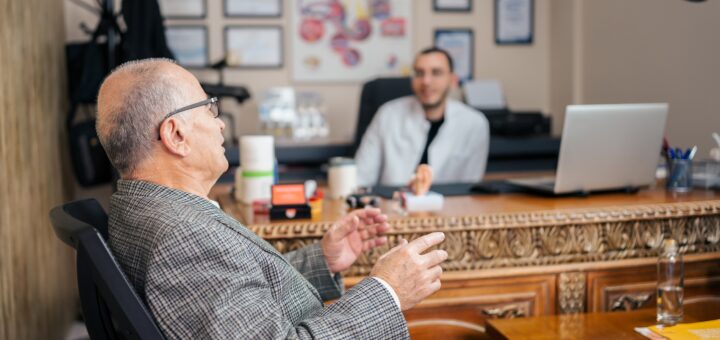 Background of a wall of framed degrees and certifications. Professional in white shirt or coat sitting behind a desk talking with older man with balding head surrounded by gray hair. The older man, wearing a gray suit sits across the desk from the professional.
