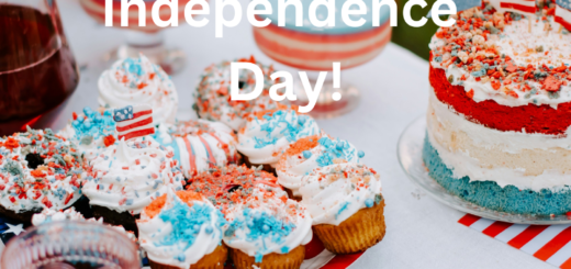 A picnic table set with cupcakes and a layer cake all decorated in red, white, and blue. Some toys flags are on the table along with a glass of red juice.