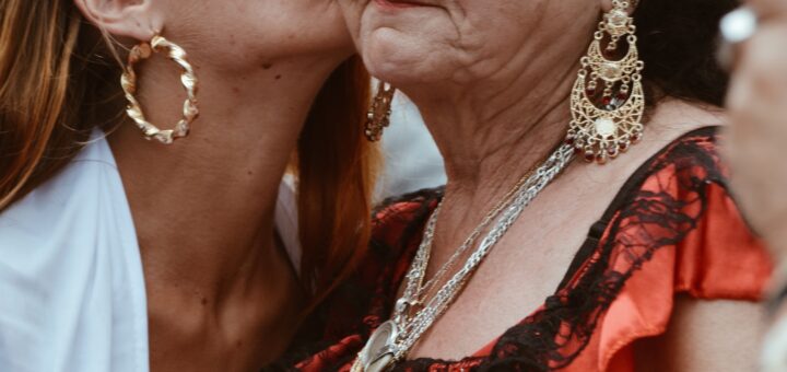 Older woman in orange and black ruffled dress looking annoyed as younger woman in white top and hoop earrings kisses her cheek