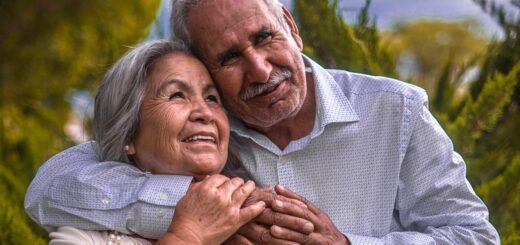 Older couple, Hispanic heritage, both with gray hair. Taller man has his arm around the woman's neck in a loving way. She is softly touching his arm and hand.