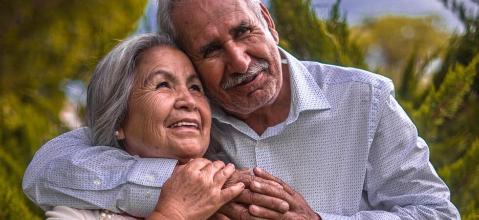 Older couple, Hispanic heritage, both with gray hair. Taller man has his arm around the woman's neck in a loving way. She is softly touching his arm and hand.