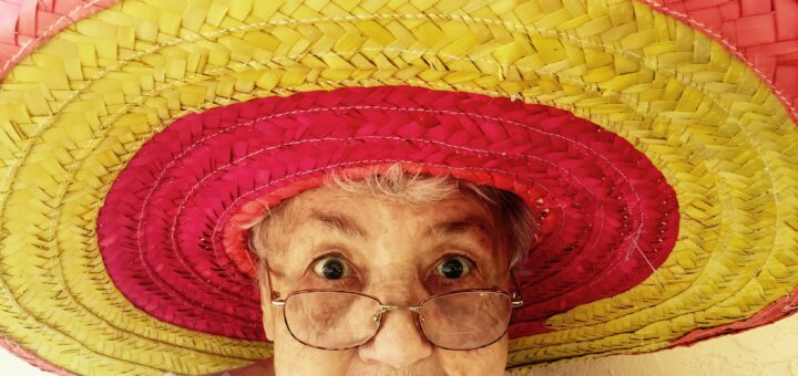 Old woman with glasses wearing a huge sombrero type straw hat colored with orange, yellow, and red circles. Her expression is bemused and slightly confused.