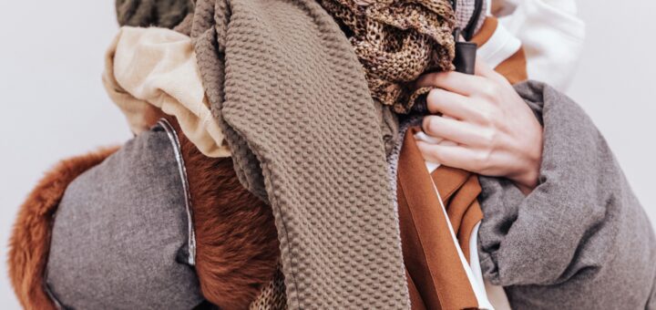 In the foreground we see a jumble of clothing, mostly in brown tones with some rush and cream. Behind the mess is a woman in a bulky coat in the same colors. We see some brownish hair on top of her head but her face is buried behind the jumbled clothing.