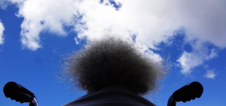 Background is a bright blue sky with clouds. An older woman with frizzy white hair is viewed from the back as she sits in a wheelchair looking up at the sky