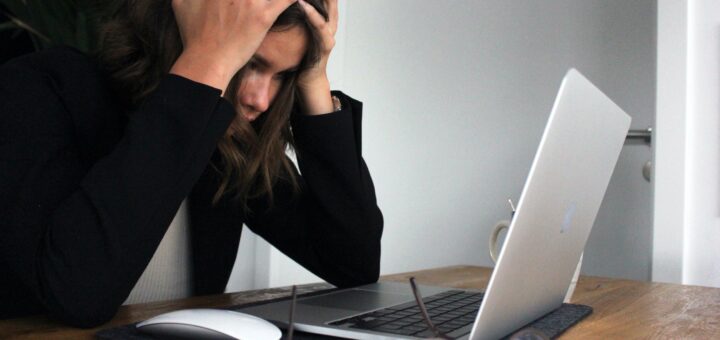 Brown haired woman sitting in front of a laptop computer, silver in color. She's holding her head with her raised hands in frustration or worry.