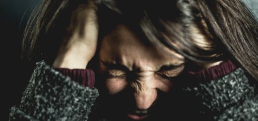 Image shows woman, face looking down with eyes squeezed shut due to frustration. Hands are on her head in her brown hair. She's wearing a black tweed sweater that shows on her forearms.