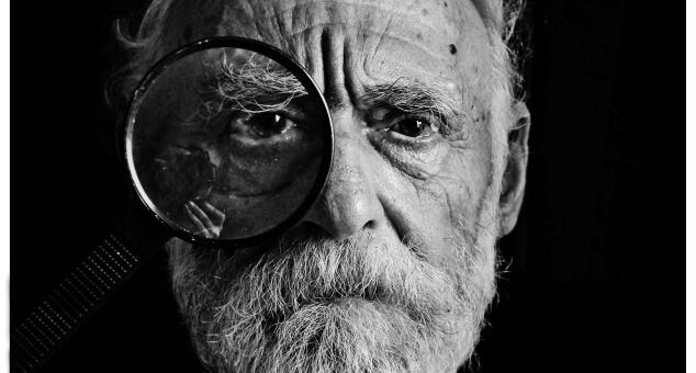 Black and white close up featuring an old man with wrinkles and a white beard looking through circle like when taking an eye test.