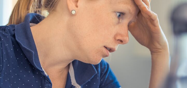 Image shows woman with light brown hair drawn into a ponytail. Her head bends forward, showing forehead in her hand. Elbow is on the table, with a water glass nearby. She wears a blue, short-sleeved shirt. The image is one of profound sadness.