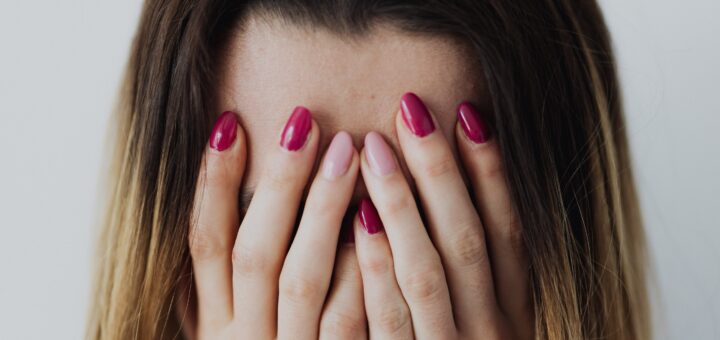 A woman covering her face with her hands as if she's crying. We see her long brown hair and both hands with long fingers and red nail polish covering her face.