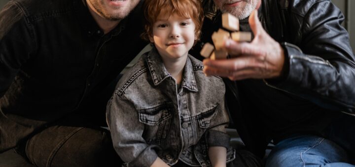 Image is family photo of three generations of males. Older man has gray hair and wears a black leather-look jacket, middle aged man with brown hair wears black cloth jacket, and child has redish-brown hair with bangs and wears a dark-colored shirt with the sleeves rolled up.