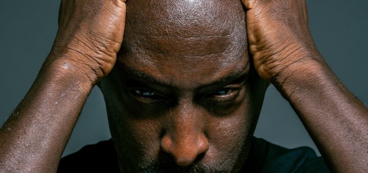 Middle aged man with dark skin and shaved head, has hands placed on top of bent head. Grief is written on his downturned face.