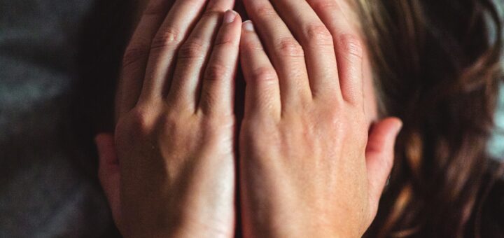 Brown haired woman lying back on bed with hands covering face in emotion and tears.
