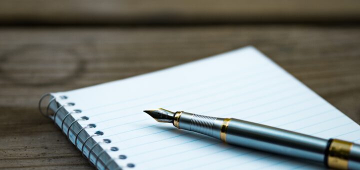 Image of a simple, spiral bound notebook laying on a rough cut table. A fountain pen sits on top of the notebook