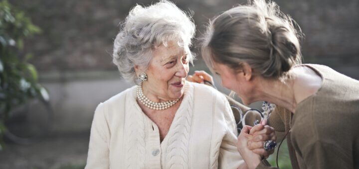 Nicely dressed older woman with gray hair. She's wearing makeup and pearls with a cream colored sweater. A younger woman with dark blonde hair in a messy bun sits by her, holding her hand and chatting with her.