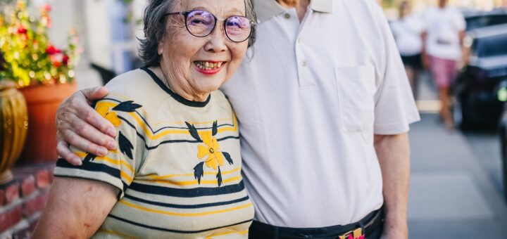 Older couple of Asian descent standing outside. Home featuring trees and flower pots. A short, gray haired woman in a light green dress smiles as her taller husband stands beside her, arm around her shoulders. He holds a a small bouquet of red and yellow freshly picked flowers in his other hand.