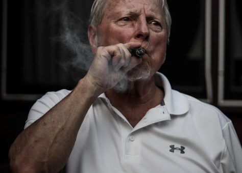 Background is dark with a feeling of being inside. Foreground shows an older man with an attitude. He's wearing a white golf shirt and smoking a cigar.