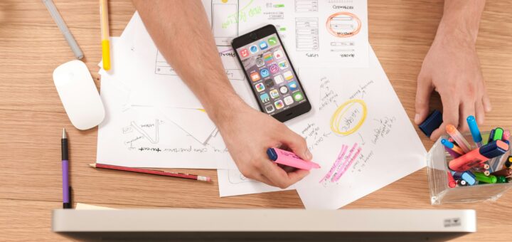 Image shows a messy desk from the tip view. Scattered about are papers, colored markers, and a smart phone. In the foreground, we view s white computer monitor from the tip edge.