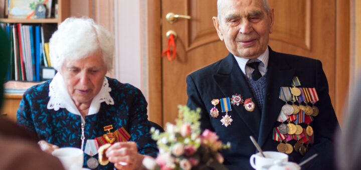 Background of a light wood interior door in a large room. Foreground is a communal dining table set with flowers, cups and other white tablewear. Sitting at the table is an older couple. The white-haired woman is wearing a blue dress with a white collar. The white-haired man wis wearing a dark blue suit with military medals pinned to the jacket.