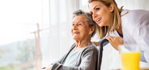 Older woman in a wheelchair wears a gray sweater. She has short, salt and pepper hair and has a appears happy. A younder woman, presumably her caregiver, is bending down to listen to the older woman. The caregiver has long brown hair, white skin, and has her hand on the wheelchair push handles.