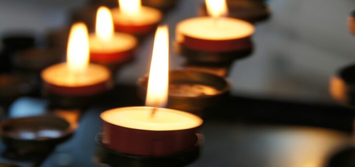 Image shows blurred dark alter or table. ON tip are rows of votive candle holders, some with lit candles and some empty. We see a total of five flames, three in back and two in the foregroud.