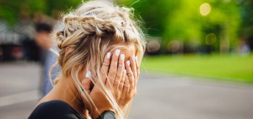 Background is parklike with trees and a parking lot. Foreground is a woman with long blonde hair done in a braid that's wrapped around her head. She's holding her face in her hands as though weeping. Her fingernails are polished in a pearl tone and she wears a watch with a wide black strap. She's standing up and she's wearing a black short-sleeved shirt.