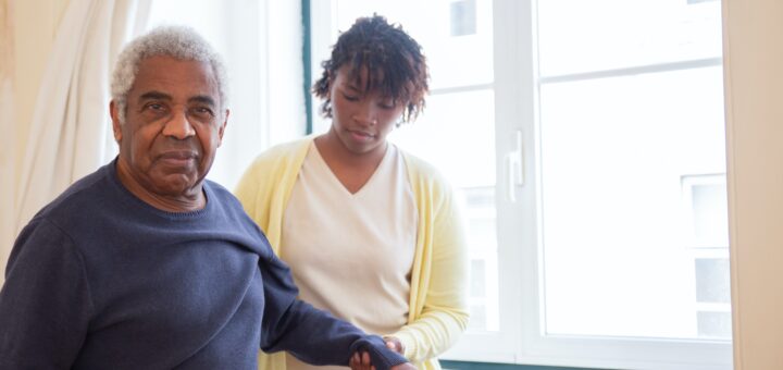 The background shows large windows in a care home. A young woman with dark hair medium toned skin is helping an older man walk down the hall. She is dressed in a white shirt with a yellow sweater. The African American man is wearing a blue sweater over a white shirt.