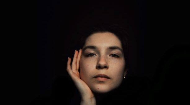 Image background is black. Foregroud is a woman's face, She has a hand resting against her cheek and her expression is pensive.