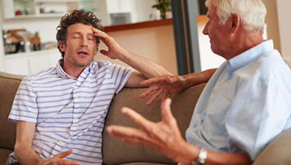 The image shows an indoor scene with an older adult and a younger man facing eacy other. The younger man has dark hair and is wearing a shirt with a white background and narrow blue stripes. He has his hand resting on his forehead and looks frustrated. The older man is shown from the profile. He's wearing a light blue shirt and has gray hair. His hand is in the foreground, open as if trying to explain something.