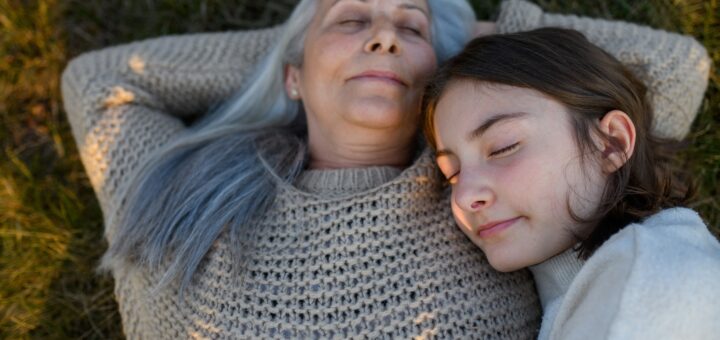 Older woman with long gray hair lies back with a content smile on her face. She's covered by a light, cream colored blanket. Younger woman with brown hair lays her head on the older womans shoulder.