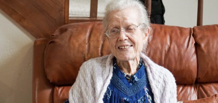 A very old woman sits on a brown leather couch. Her gray hair is pulled back, and she wears rimless glasses. Her dress is dark blue and she's wearing a cream-colored knit shawl. Her expression is pleasant.