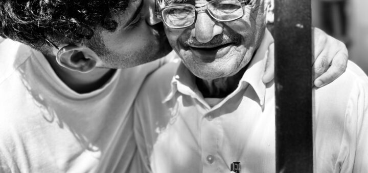 Balck and white image of a younger person with curly dark hair and wearing a white shirt bending over to kiss an older man on the cheek. The older man is smiling with please. He wears a white shirt and glasses, and is mostly bald.