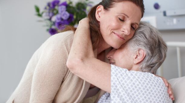 The background shows a white wall with a hanging of purple flowers. Foreground shows an older, gray haired woman in a hospital gown hugging a younger woman. The younger woman has long dark hair and is wearing a beige sweater.