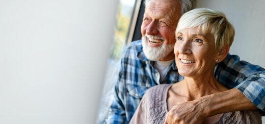 Image shows a curtained background with a happy older couple in the foreground. T he man stands with his arm around the woman's shoulders. He has gray hair and a white beard and wears a blue and white plaid shirt. The smiling woman has short white hair and is wearing a scoop-necked blouse or gown in a small blue print on a white background.