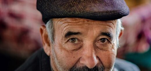The image shows a closeup of an older man with a gray beard and a salt and pepper mustache. He's wearing a dark purple porkpie hat and a gray doat. His expression shows subtle humor mixed with defiance.