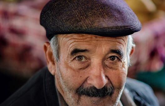 The image shows a closeup of an older man with a gray beard and a salt and pepper mustache. He's wearing a dark purple porkpie hat and a gray doat. His expression shows subtle humor mixed with defiance.