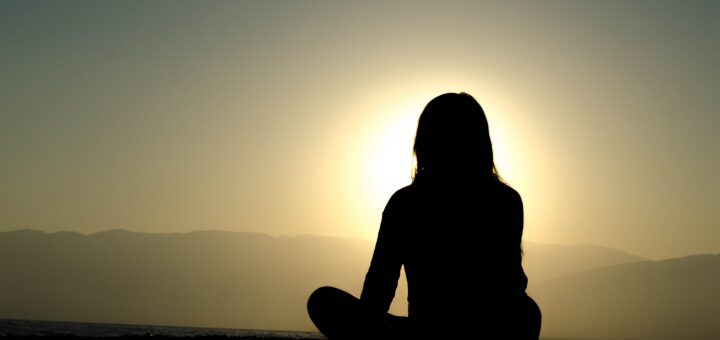 Background is a rising sun over hills. Foreground shows a back view of a siloouetted woman seated in a cross-legged yoga pose, arms in front. She's gazing toward the horizon.
