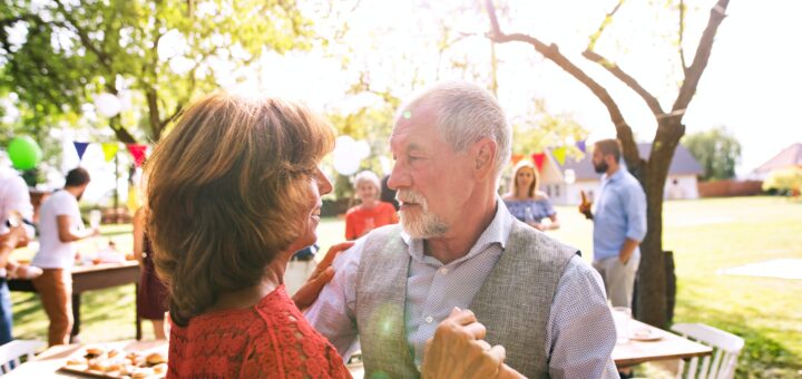 An adult daughter with brown hair and wearing a red dress is showing dancing with an older gray-haired man, likely her father. The background shows lovely spring trees and a gathering that could be in a park.