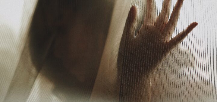 Image shows a female silhouette peering between window sheers.. Her hand is on the glass and she looks like she's wishing for help