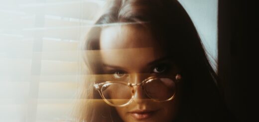 A window with a sheer covering shows a blurred image of a woman looking out. She's got long brown hair and is looking over large glasses.